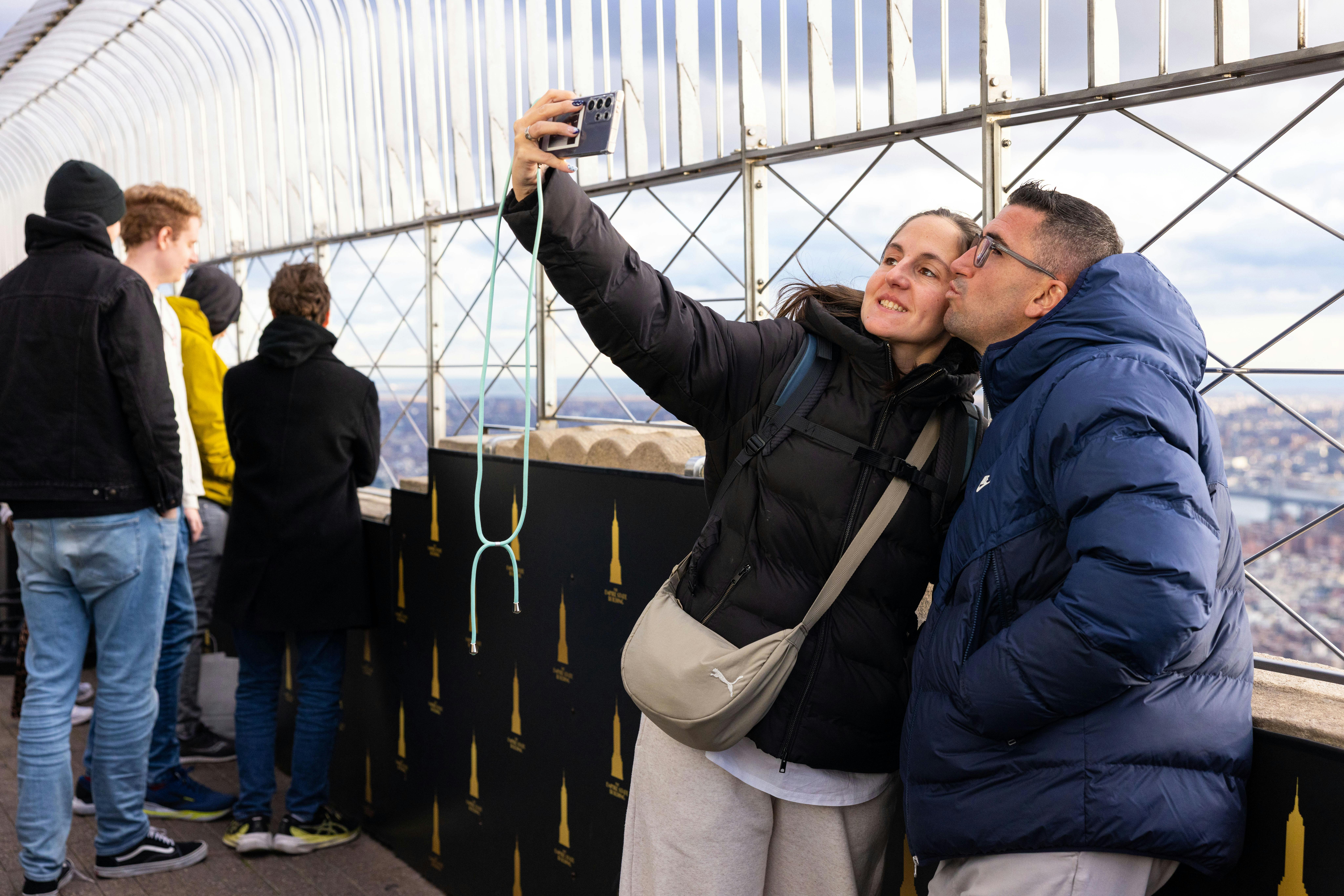 Two people in warm jackets take a selfie at an elevated outdoor location with a metal railing and cityscape in the background.