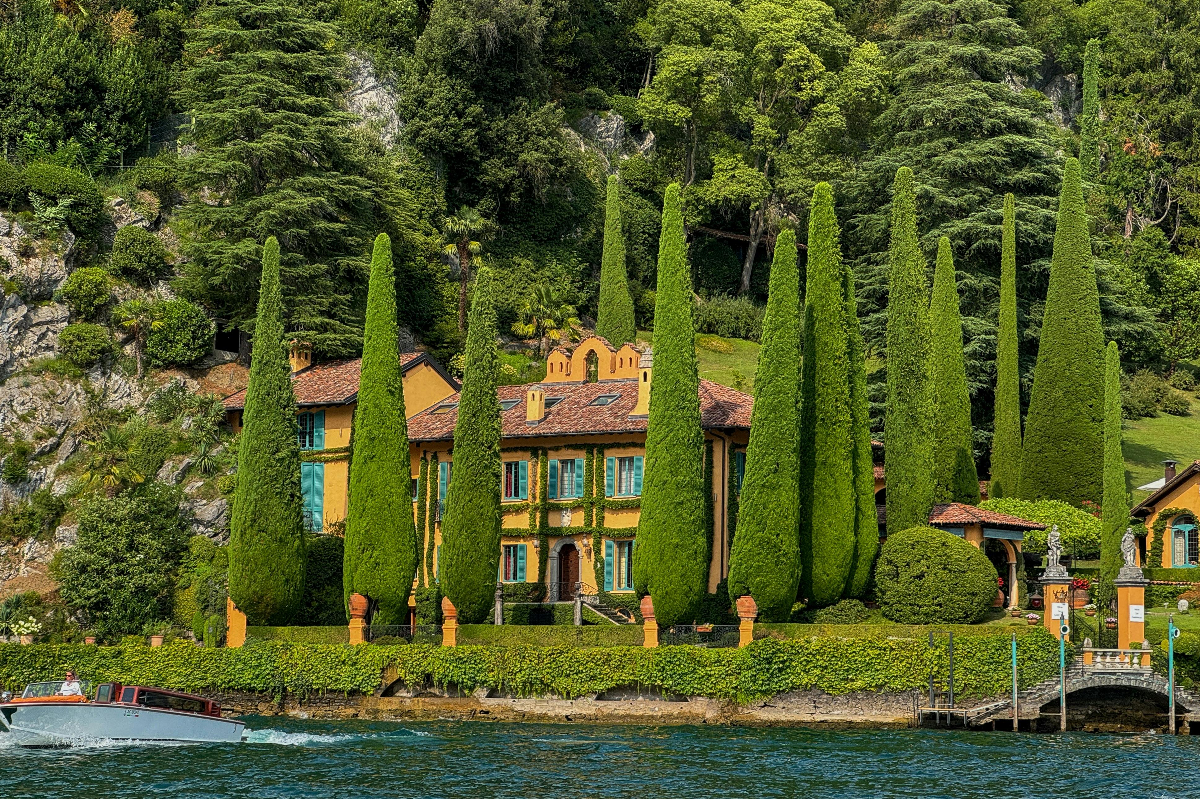 A villa with yellow walls and green shutters surrounded by tall cypress trees, near water, with a forested hillside behind.