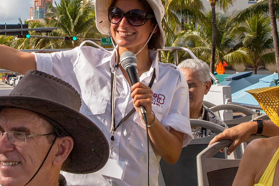 A tour guide speaks to a group of people on an open-top bus on a sunny day, surrounded by palm trees and buildings.