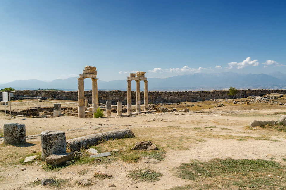 Ancient stone pillars and ruins in a barren landscape with distant mountains under a clear blue sky.