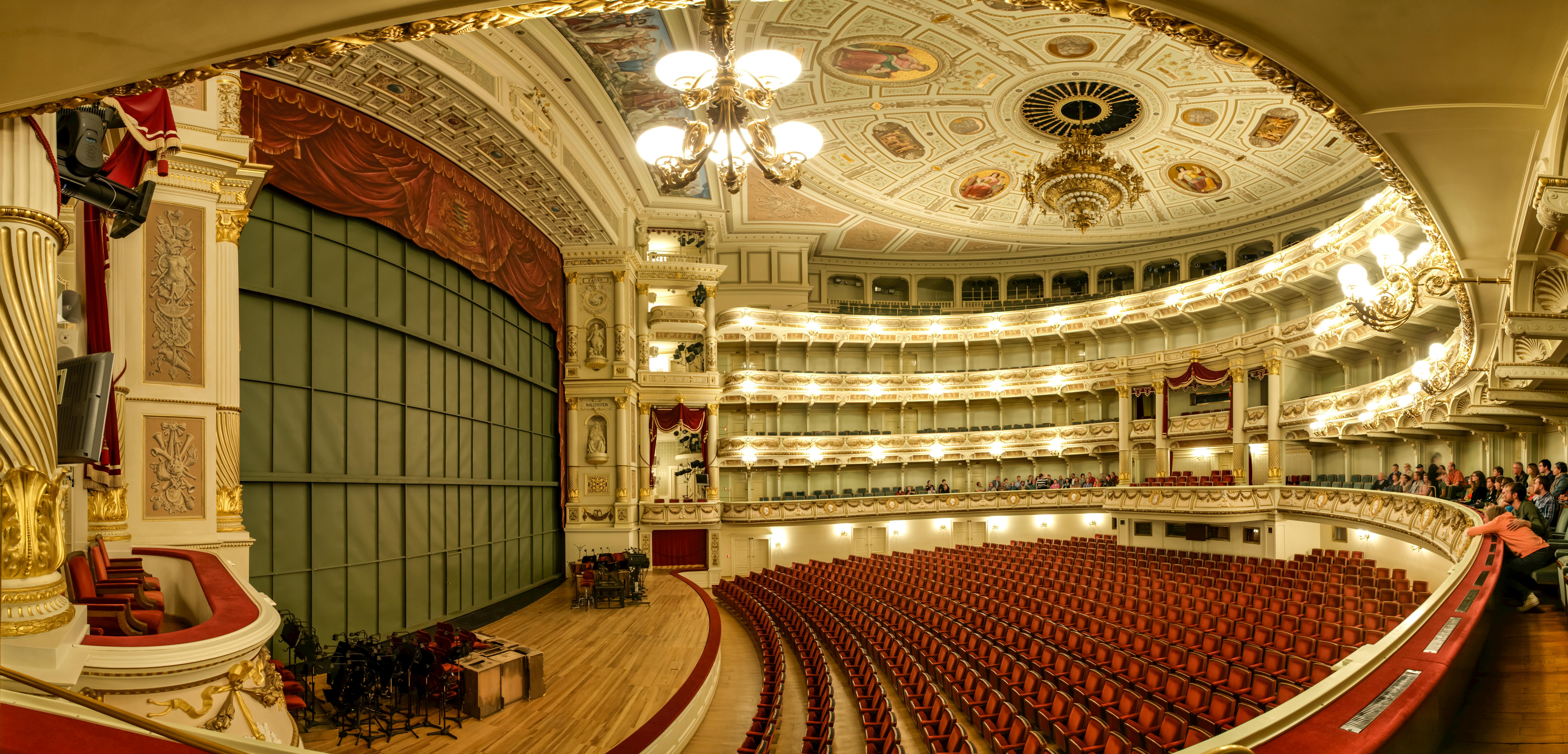 Opulent theater with ornate, decorated ceiling, chandelier, balconies, red chairs, and a stage with a red curtain.