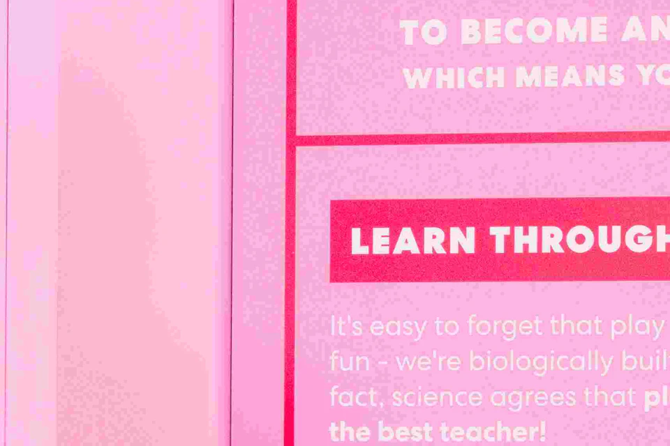 Two women interact in a bright pink room with mission instructions on the wall. A man stands nearby, partially out of frame.