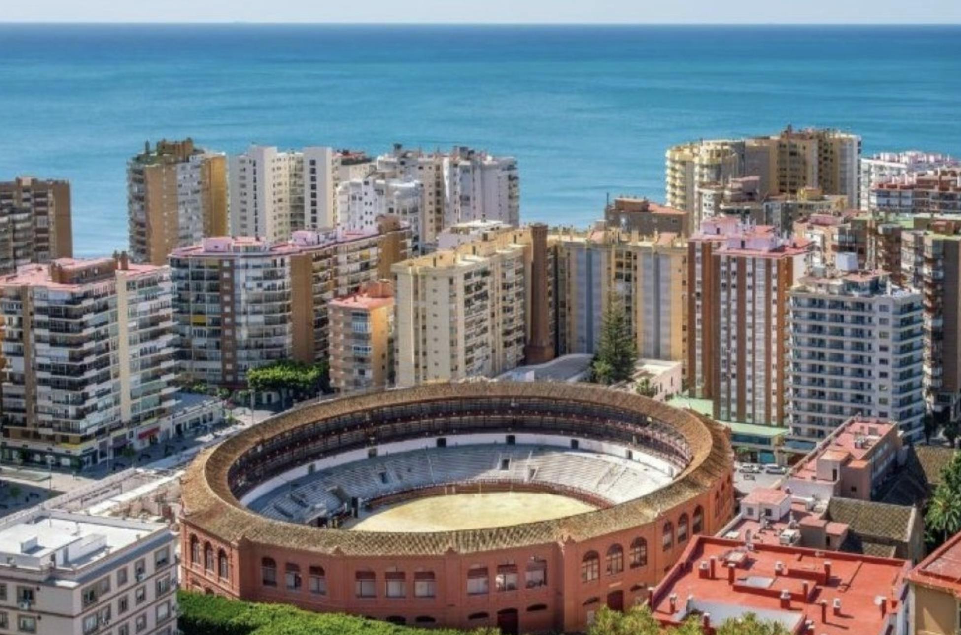 Aerial view of a coastal city featuring a round, red-brick arena surrounded by tall residential buildings with the sea in the background.