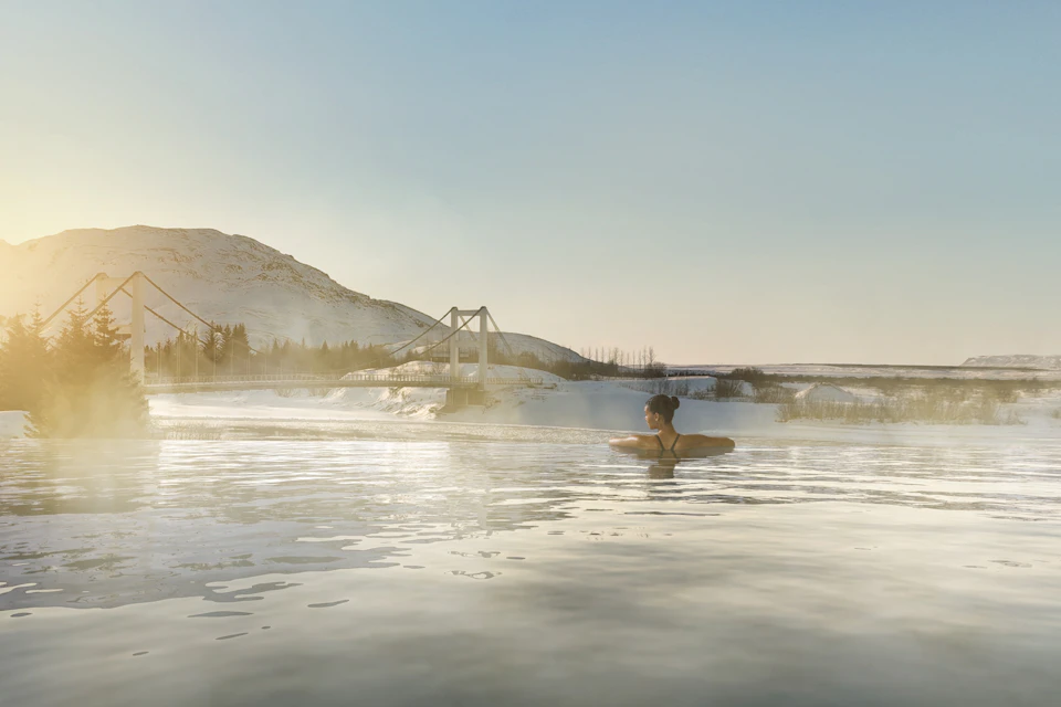A person relaxes in a steaming outdoor pool with a snowy landscape, a bridge, and mountains in the background under a clear sky.