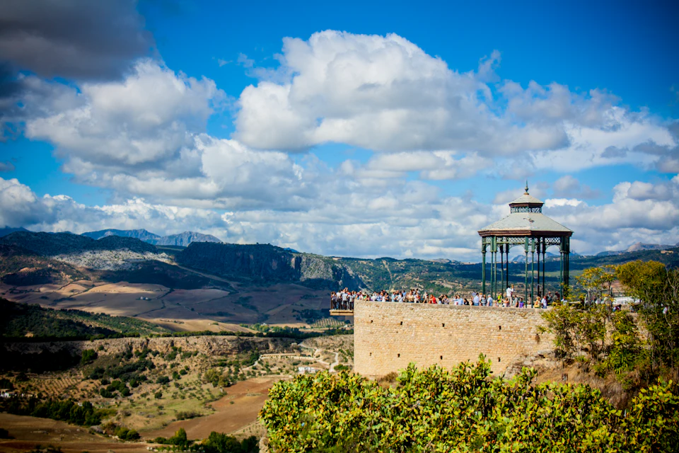 People gather at a scenic viewpoint with a gazebo, overlooking a vast landscape of hills and valleys under a partly cloudy sky.