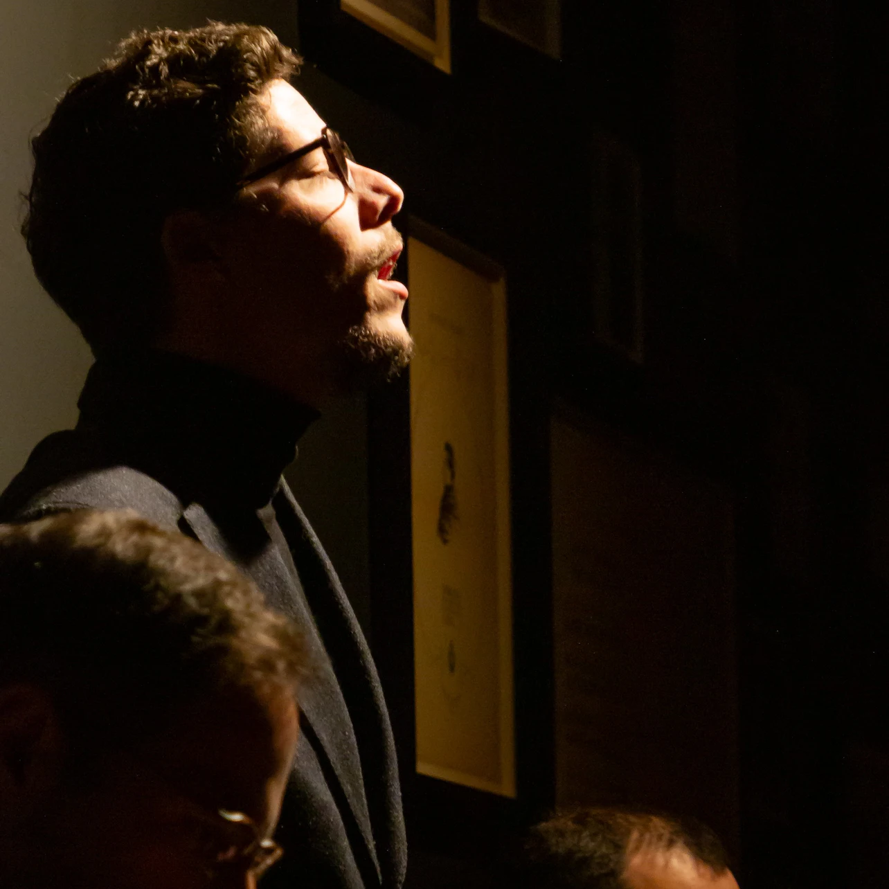 A man with glasses and a beard, wearing a dark turtleneck, is illuminated dramatically against a dark background with framed pictures.