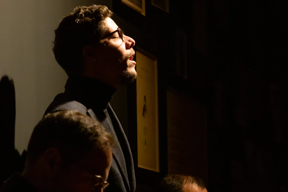A man with glasses and a beard, wearing a dark turtleneck, is illuminated dramatically against a dark background with framed pictures.