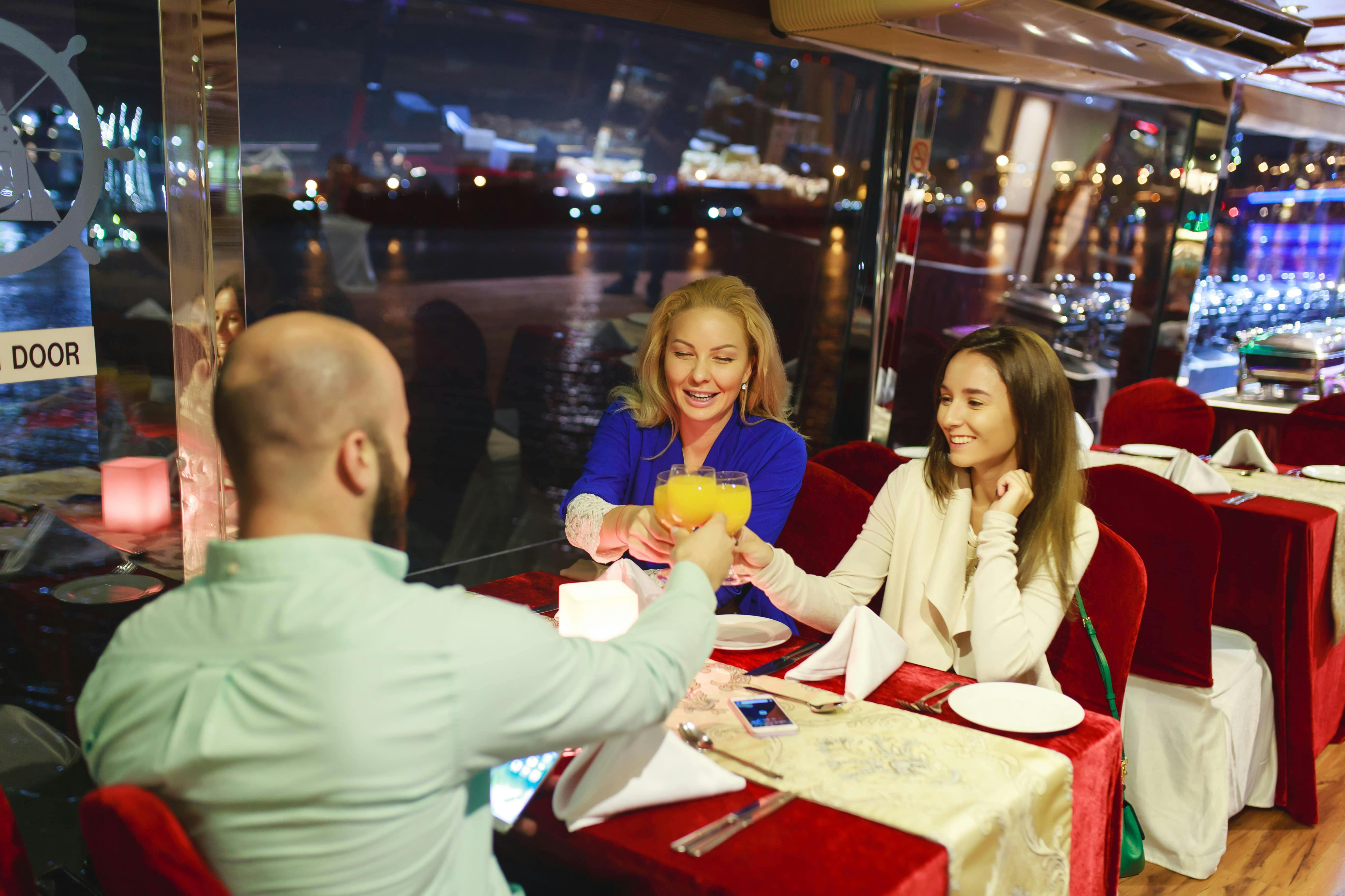 Three people toasting with drinks at a restaurant table by a window overlooking a nighttime cityscape.