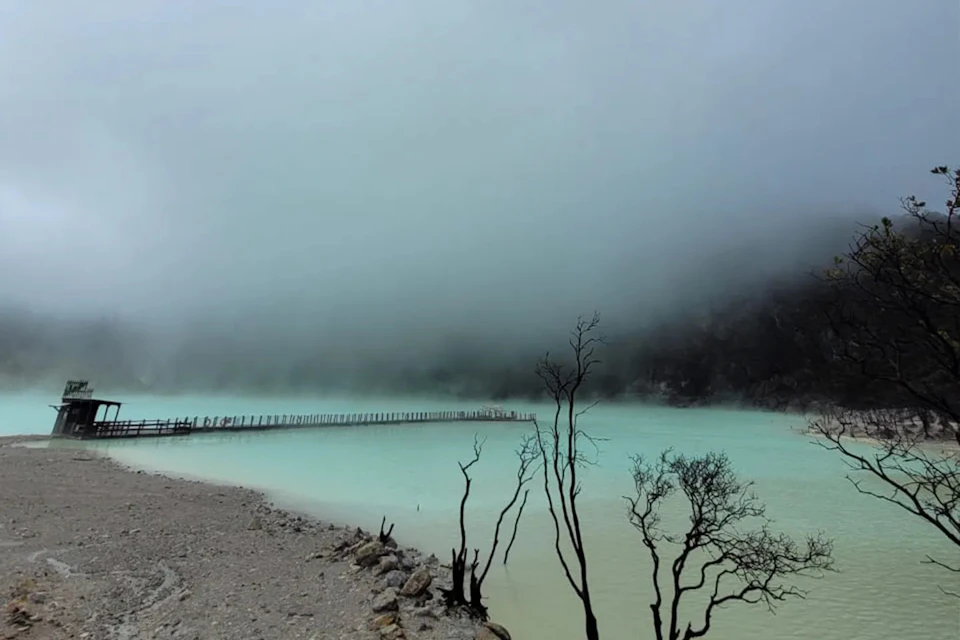 A misty lake with turquoise water, a wooden walkway crossing it, and sparse leafless trees in the foreground.