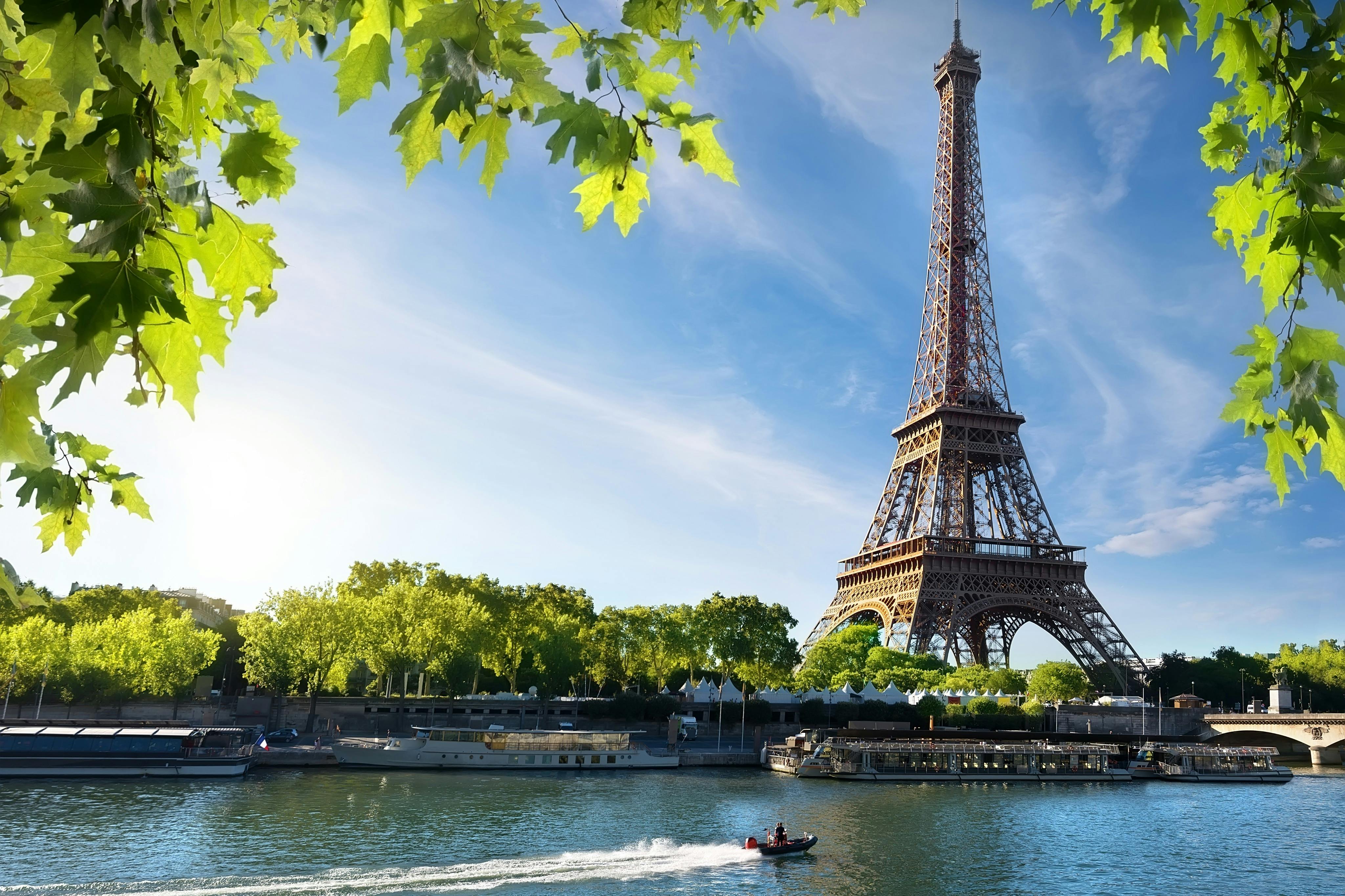 La Torre Eiffel vista dall'altra parte della Senna, con barche sull'acqua e alberi verdi sotto un cielo soleggiato.