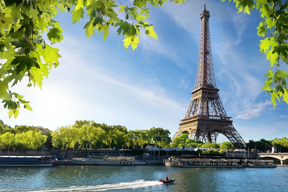 The Eiffel Tower viewed from across the Seine River, with boats on the water and green trees under a sunny sky.