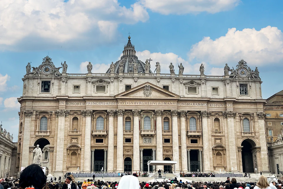 St. Peter's Basilica with a crowd gathered in the foreground under a blue sky with clouds.