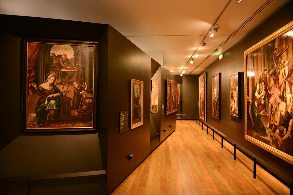 A museum hallway features several framed paintings on dark walls with wooden floors and overhead lighting.