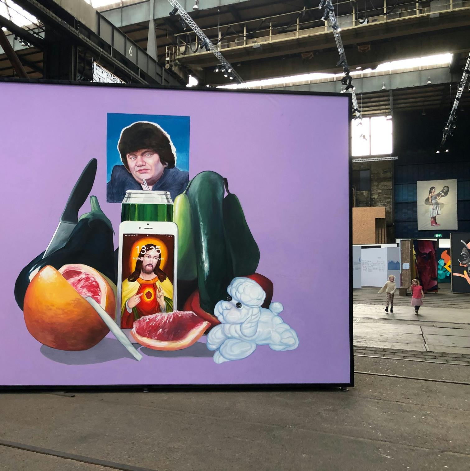 산업 공간에서 과일, 촛불, 한 남자의 초상이 담긴 대형 작품. 사람들이 작은 작품을 배경으로 감상하고 있습니다.