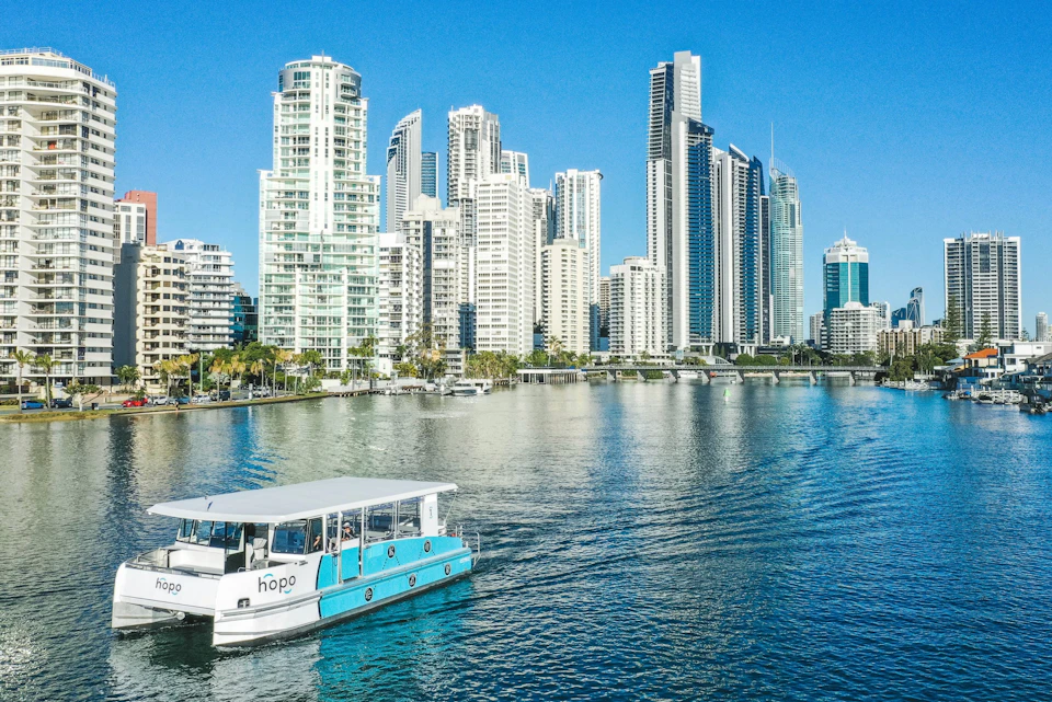 A ferry on a calm river with a skyline of modern high-rise buildings under a clear blue sky.