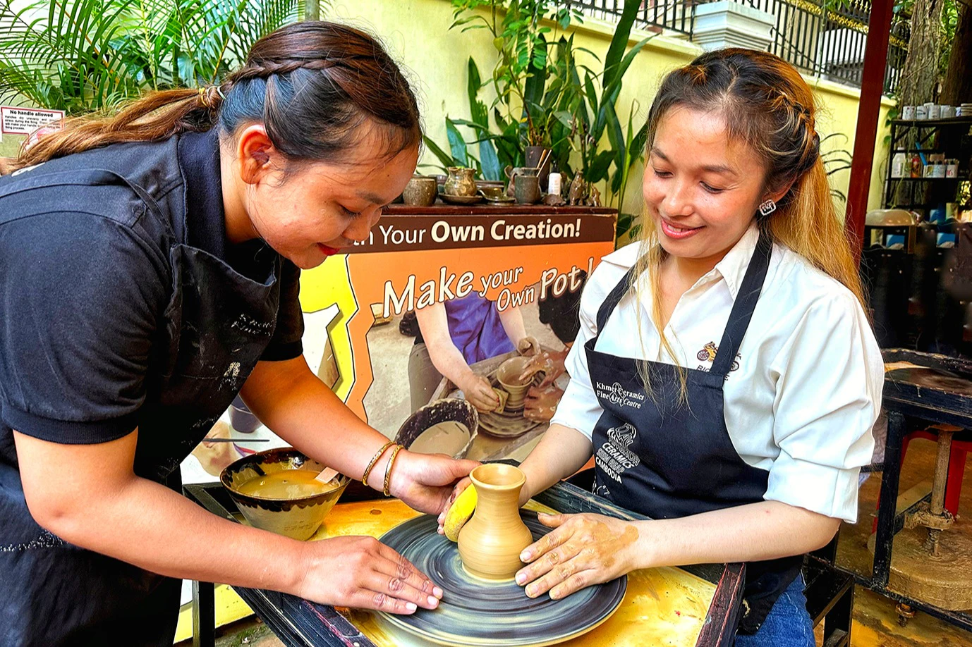A woman wearing an apron shapes clay on a pottery wheel, assisted by another person, with a sign promoting pottery in the background.
