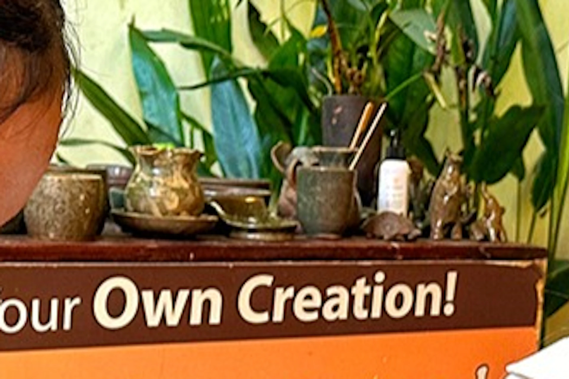 A woman wearing an apron shapes clay on a pottery wheel, assisted by another person, with a sign promoting pottery in the background.