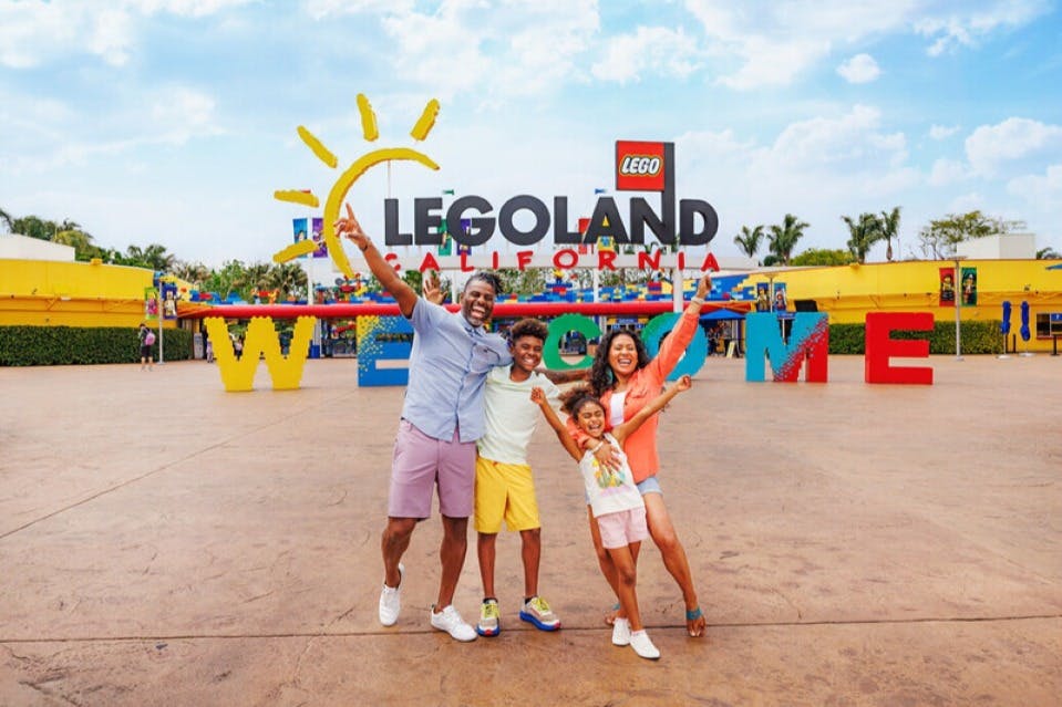 En glad familj poserar framför den färgglada välkomstskylten LEGOLAND California en ljus dag.