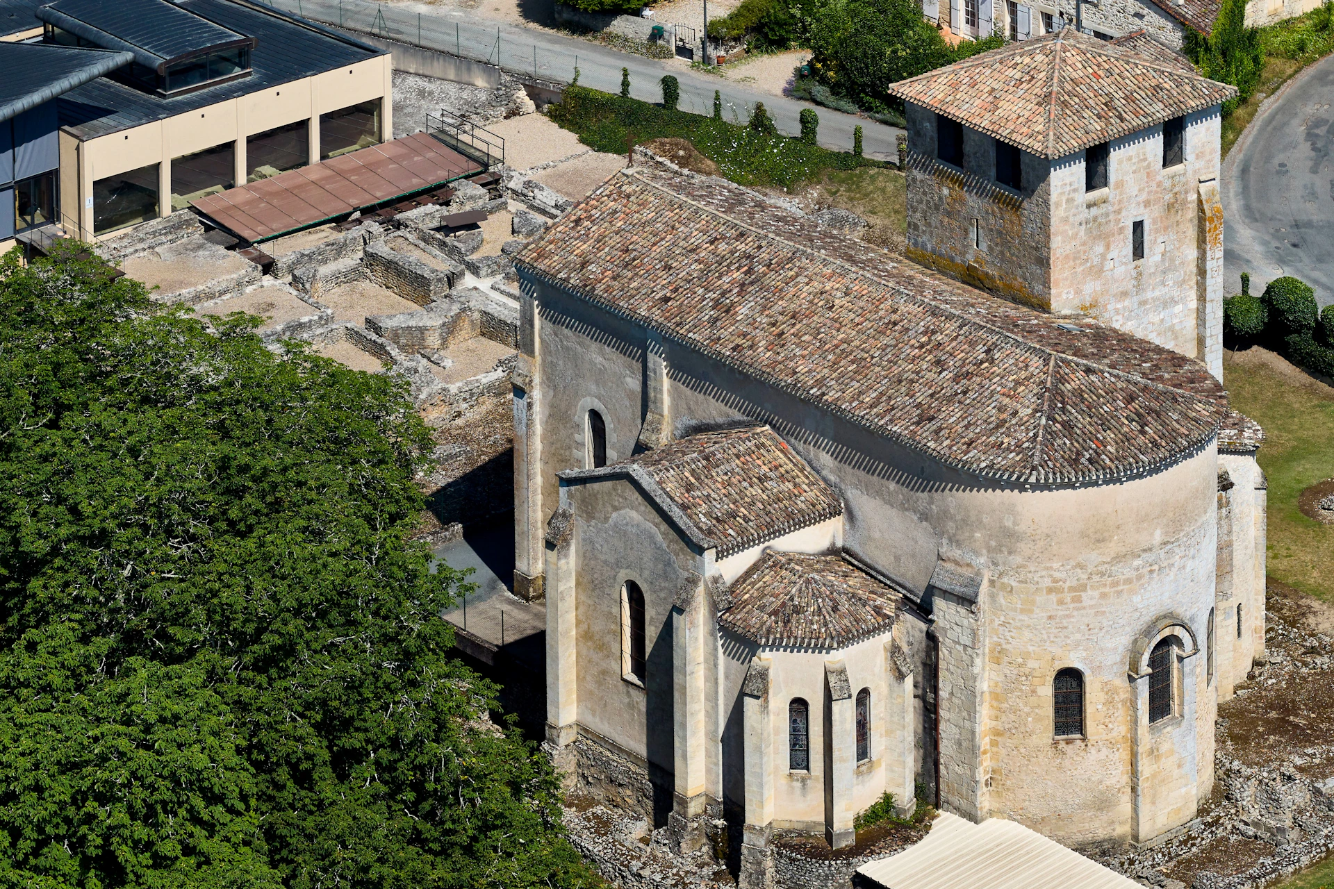Aerial view of an old stone church with a tiled roof, adjacent ruins, and nearby buildings and greenery.