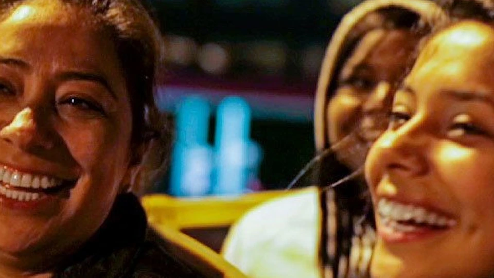 Two people smiling and laughing on an open-top bus at night, with a cityscape backdrop.