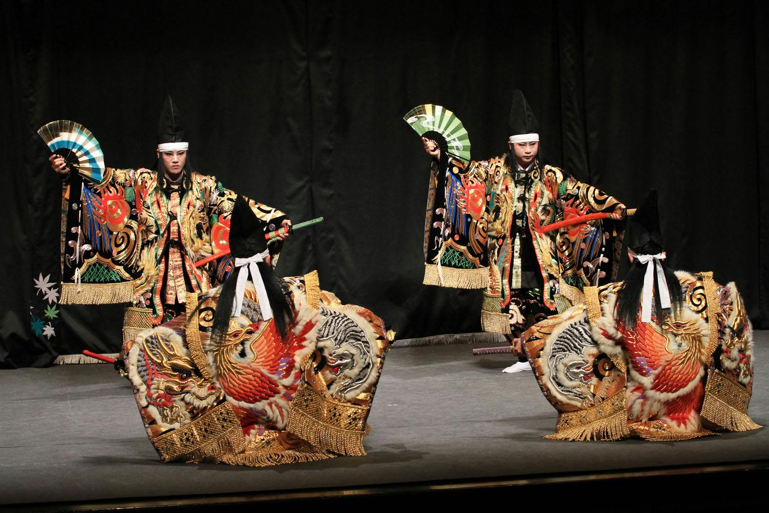 Performers in elaborate traditional costumes with ornate embroidery, holding decorative fans on a dimly lit stage.