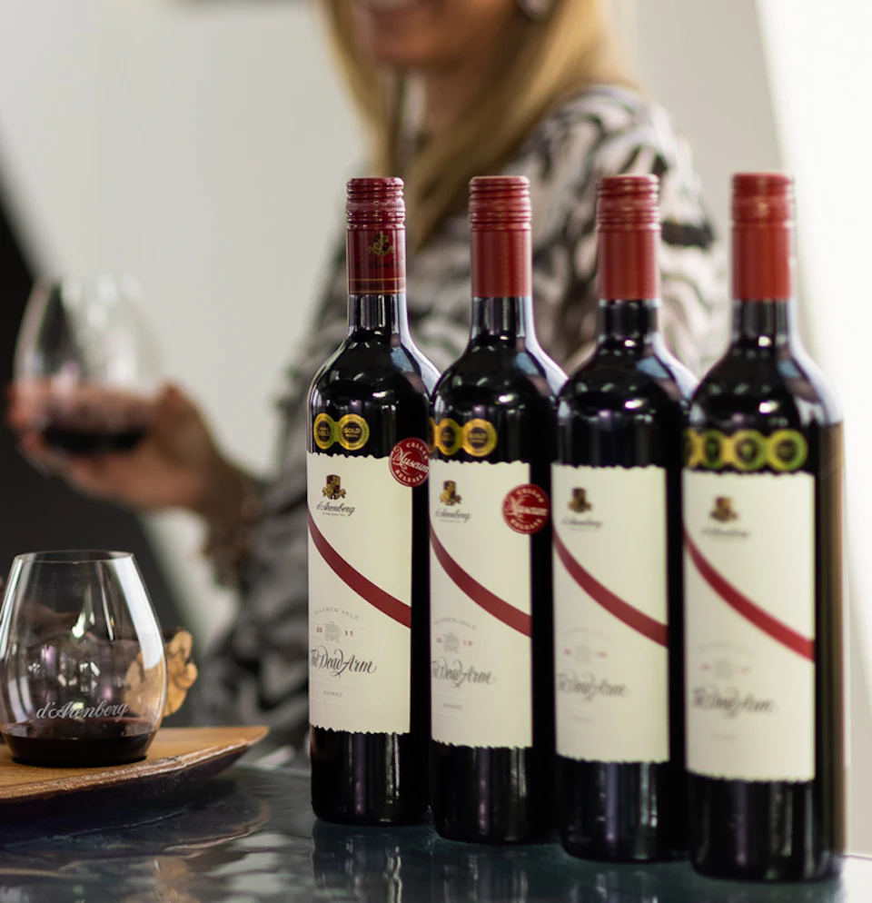 Four bottles of red wine on a counter with a person holding a glass of wine in the background.