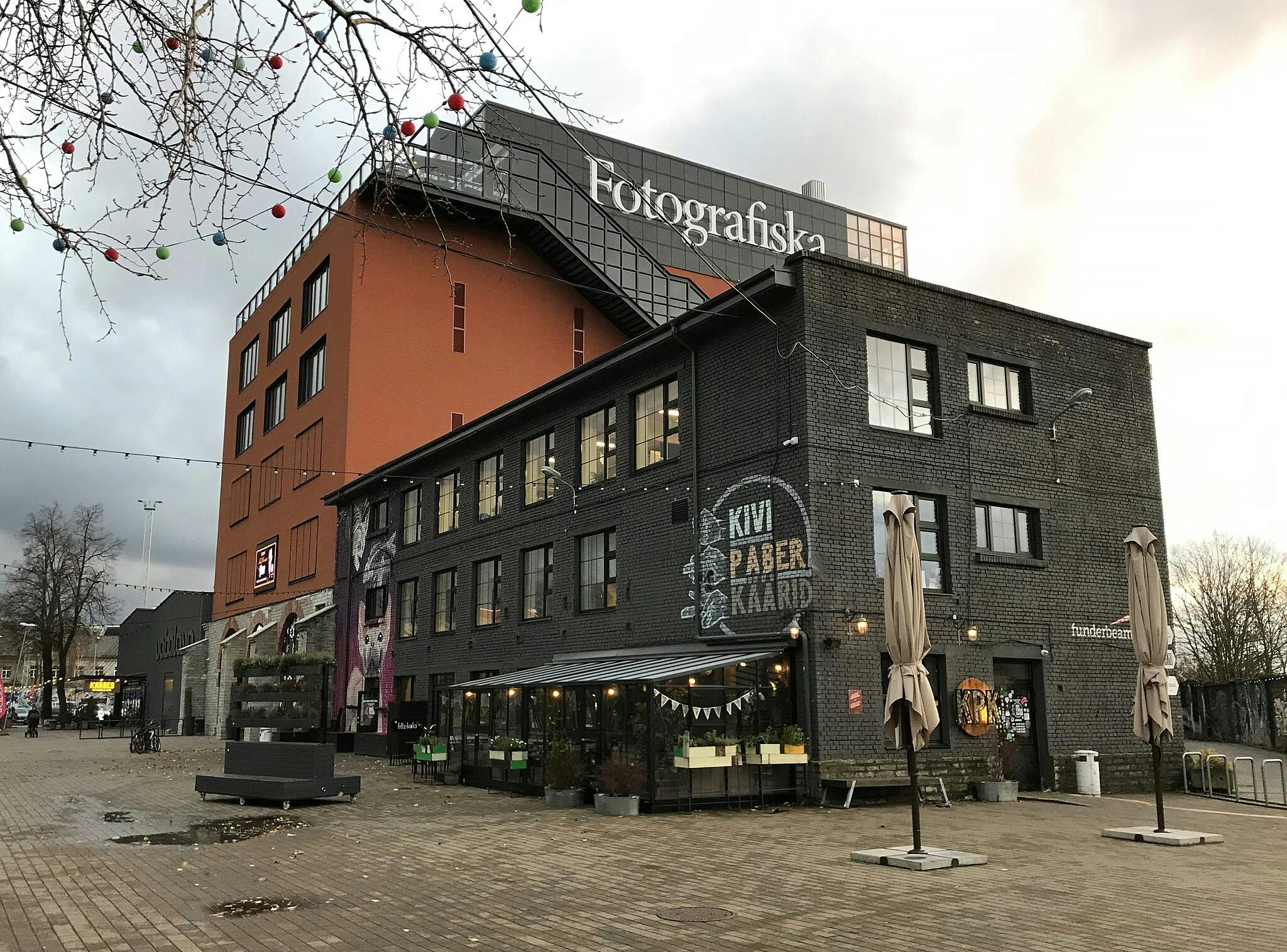 A city street scene with a modern building labeled "Fotografiska," adjacent to a black brick building with "Kivi Paber Käärid" signage.