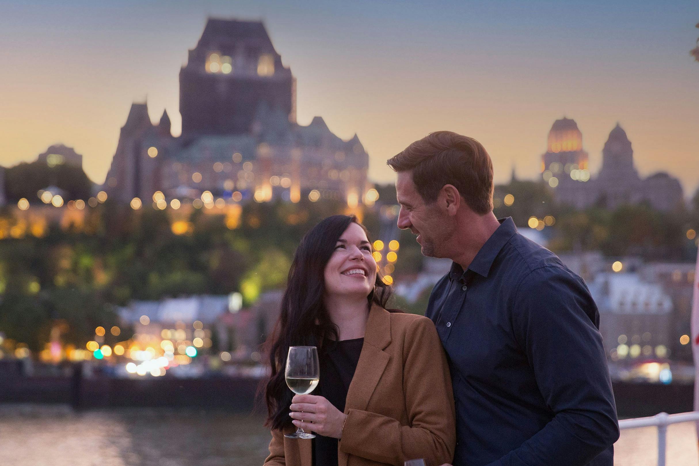 Un couple se sourit en tenant un verre de vin, avec un paysage urbain et des bâtiments illuminés en arrière-plan.