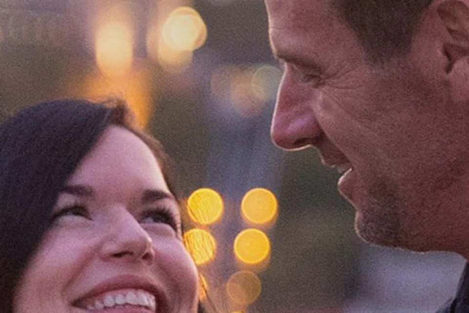 A couple smiling at each other while holding a glass of wine, with a cityscape and illuminated buildings in the background.