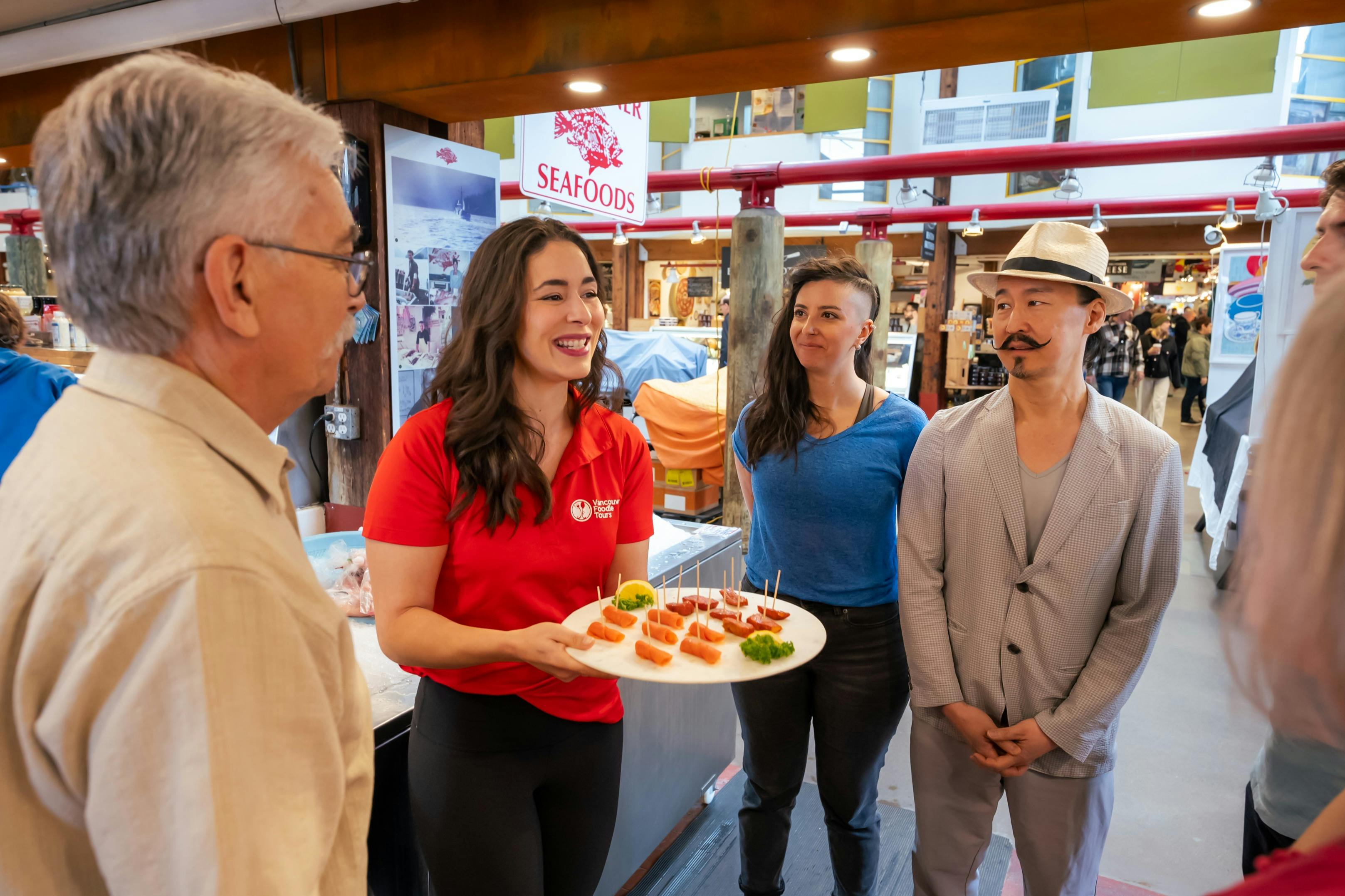 빨간 셔츠를 입은 여성이 해산물 시장에서 세 사람에게 음식 샘플을 제공합니다.
