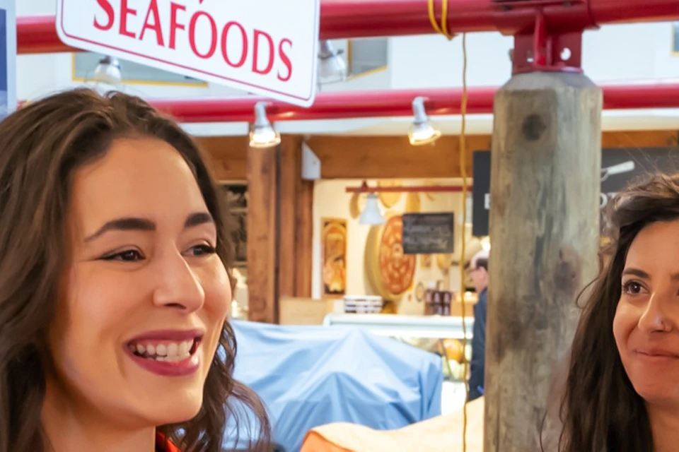 A woman in a red shirt offers a plate of food samples to three people in a seafood market.