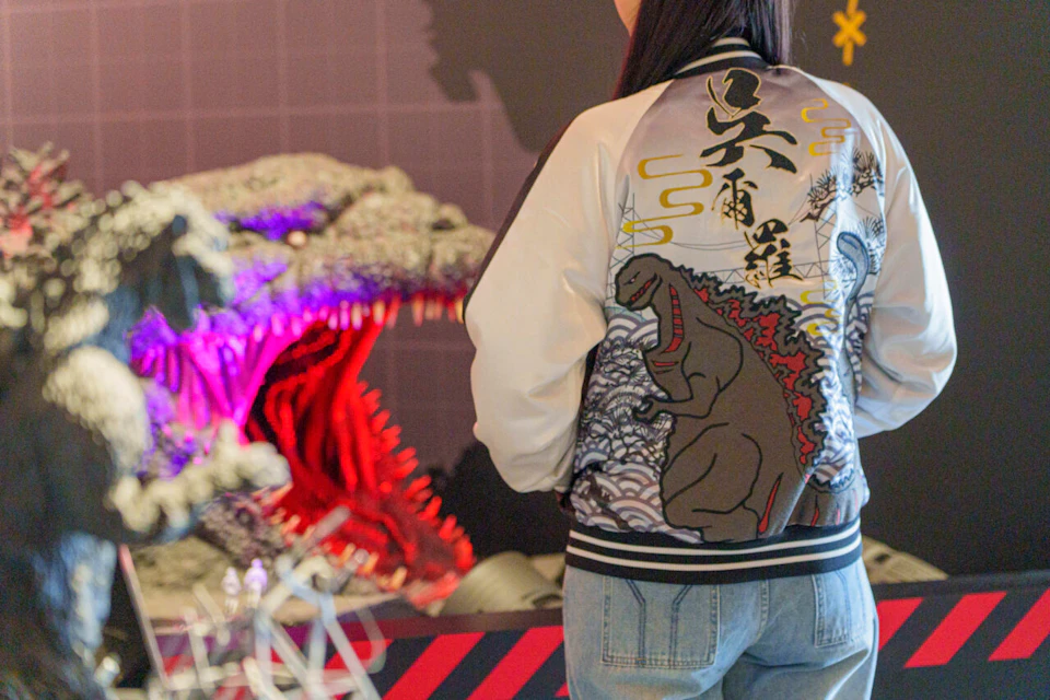 A person wearing a jacket with a Godzilla design stands in front of a display featuring a large Godzilla model with red lighting.