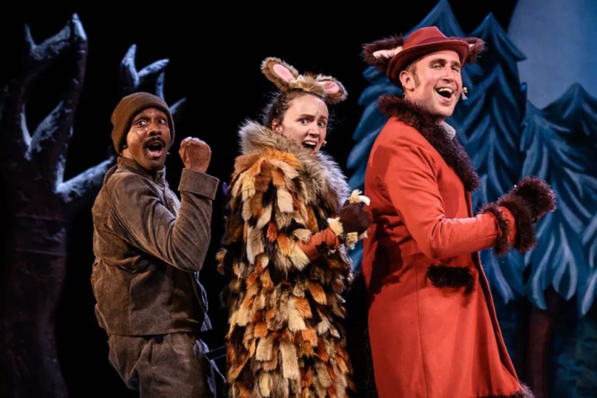 Dva herci na jevišti ve zvířecích kostýmech, jeden v chlupatém outfitu s ušima a druhý v červeném kabátě s kloboukem a kožešinovým lemováním.
