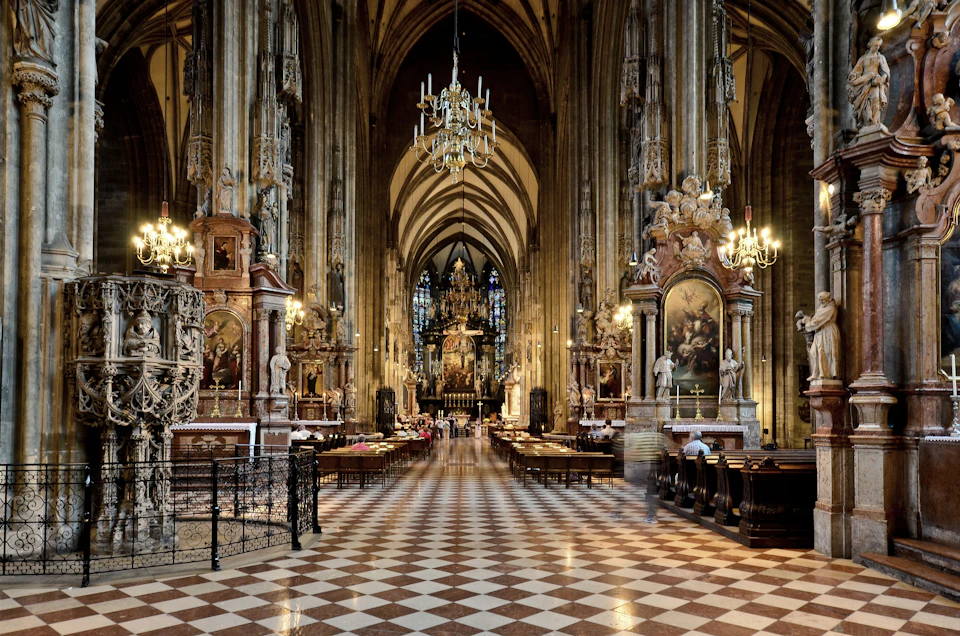Interior of a grand cathedral with high vaulted ceilings, chandeliers, stained glass, and ornate religious sculptures.