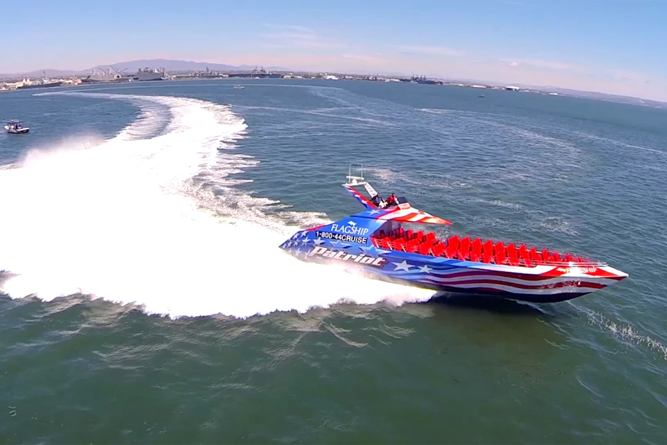 A speedboat decorated in the American flag colors cuts through the water, creating a large wake against a backdrop of distant ships and city.