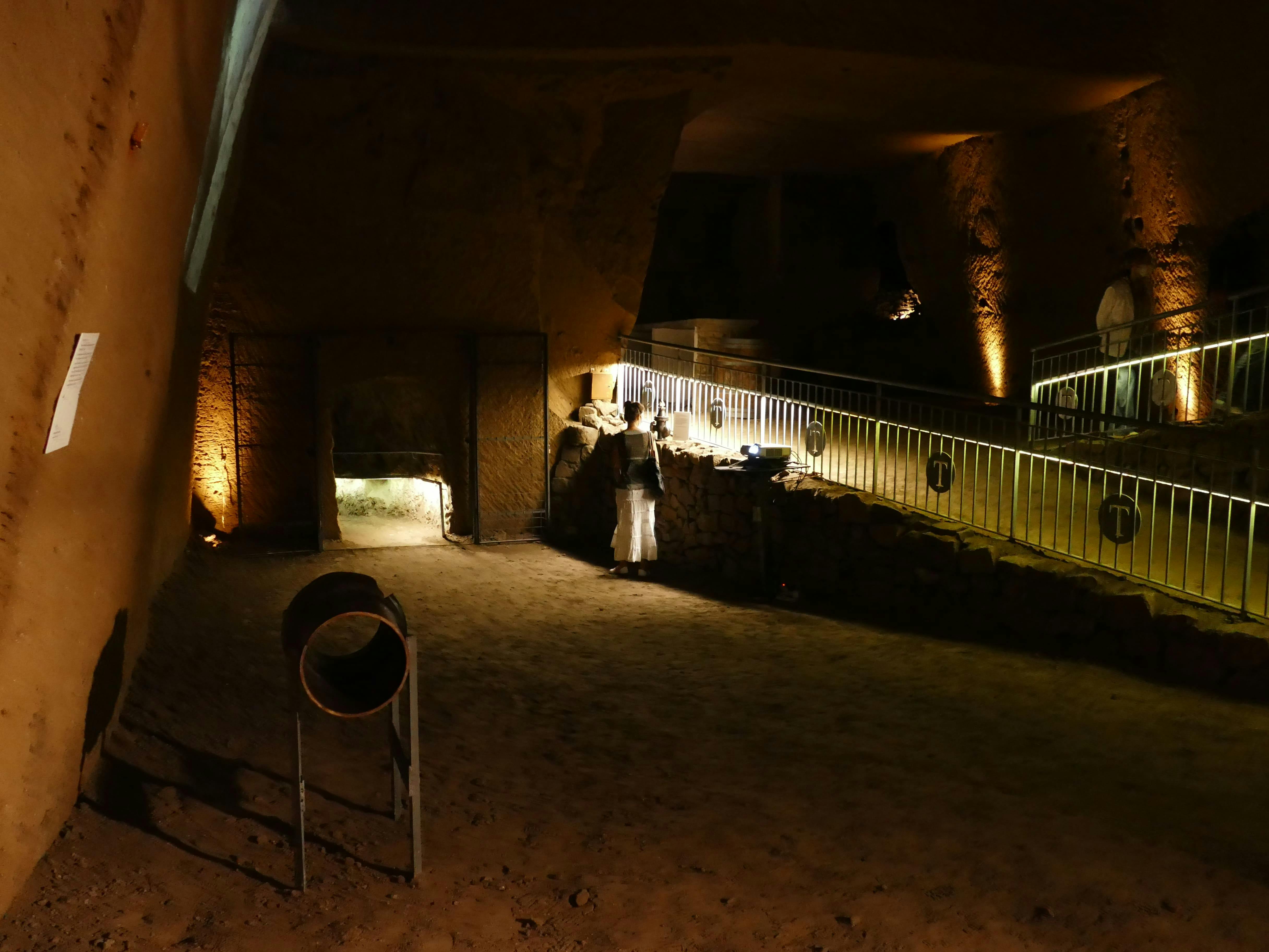 A person in a dimly lit underground cavern, standing beneath a stone arch. Railings and lights line a pathway above.
