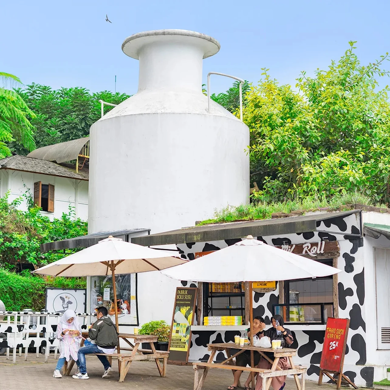 People sitting at picnic tables under umbrellas outside a cow-themed food stand with greenery and a white structure nearby.