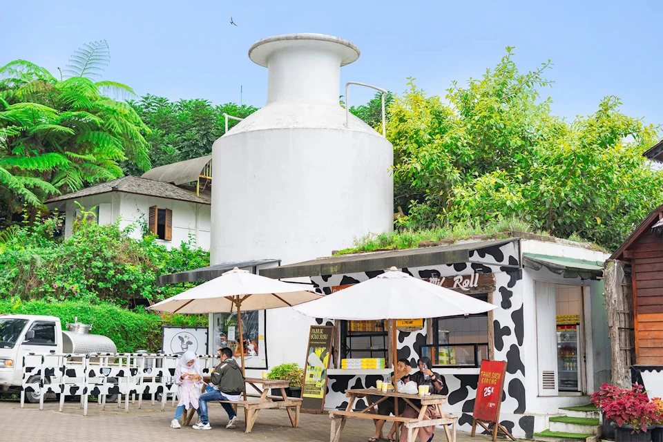 A small outdoor cafe with cow-patterned walls, wooden tables, and umbrellas, surrounded by greenery and a white silo structure.
