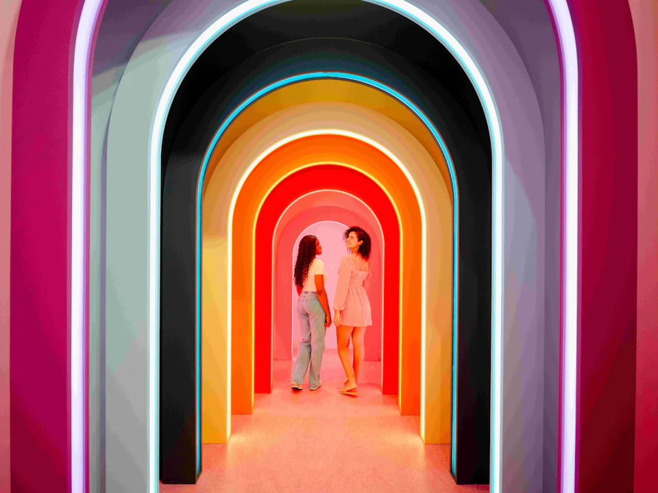 Two women standing under multicolored illuminated arches in a hallway, facing each other.