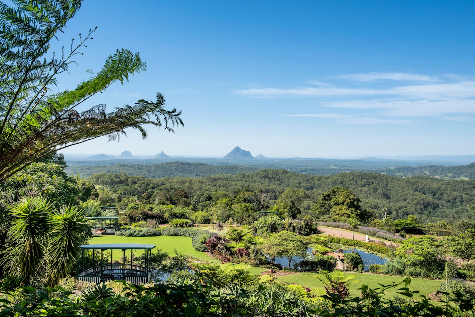 A lush garden with a pond and gazebo overlooks a forested landscape and distant mountains under a clear blue sky.