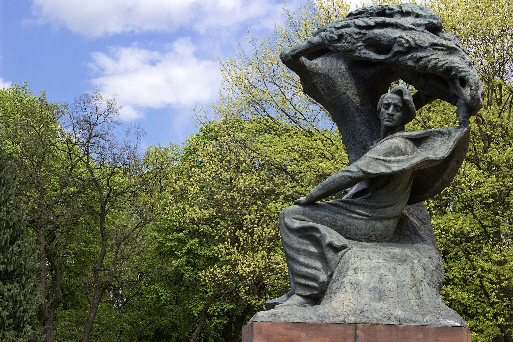 울창한 푸른 나무와 구름이 낀 푸른 하늘로 둘러싸인 공원에 망토가 흐르는 앉아있는 모습의 청동 동상.