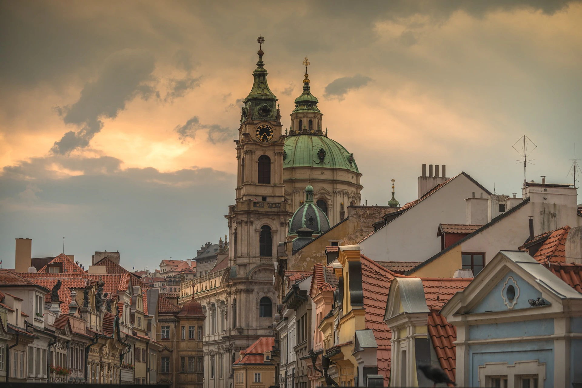 Historic cityscape with St. Nicholas Church towers at sunset, surrounded by old buildings with red-tiled roofs.