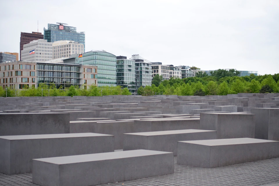 A field of gray rectangular blocks in varying heights, with modern buildings and trees in the background.