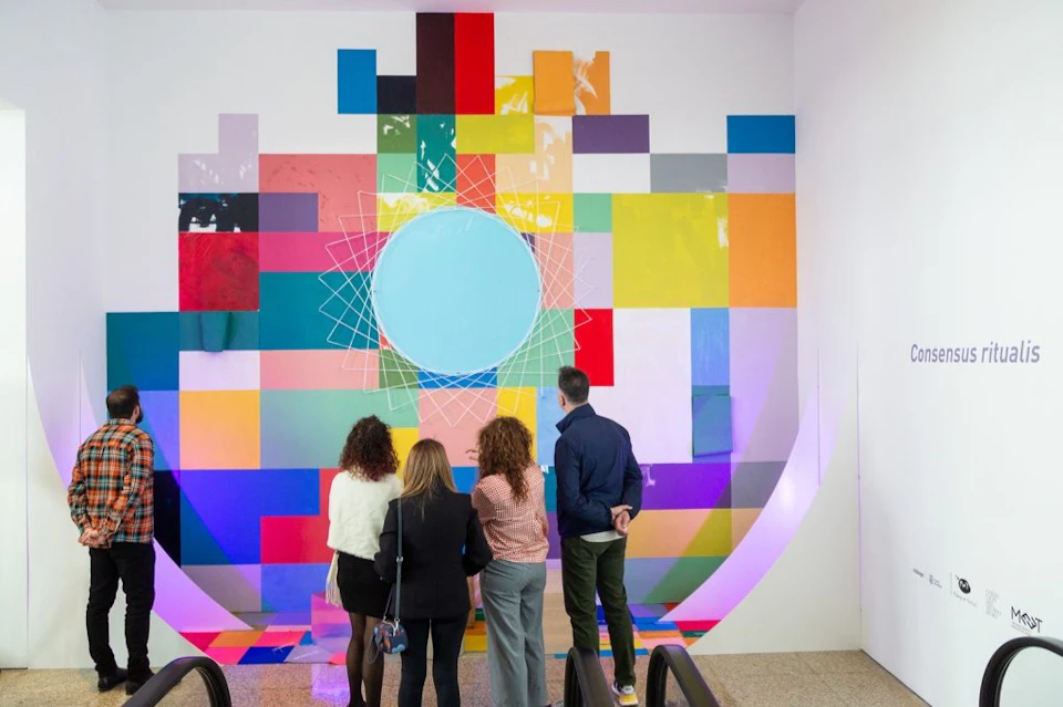Four people are standing in front of a colourful abstract art installation featuring geometric patterns, looking at the display.