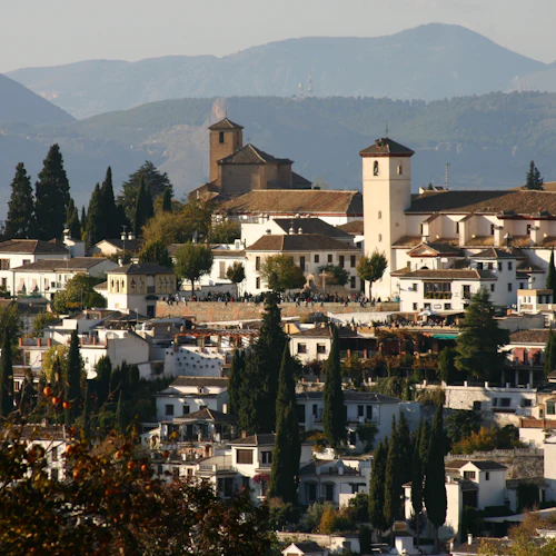 En bergsby med vita byggnader och ett framträdande kyrktorn, omgivet av träd, med berg i bakgrunden.
