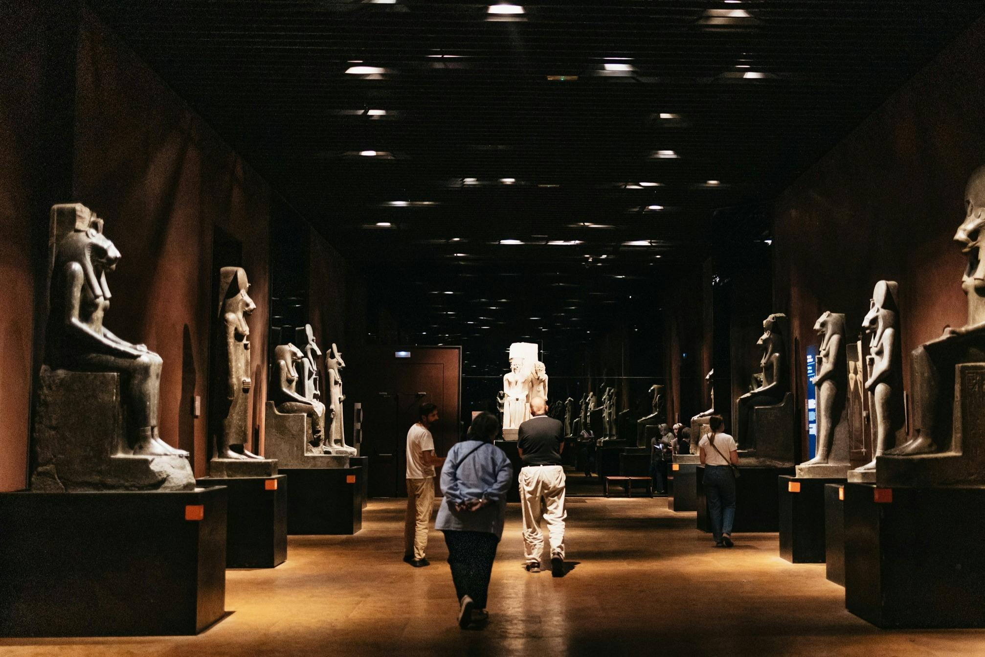People walking through a dimly lit museum gallery with statues displayed on pedestals along the walls.