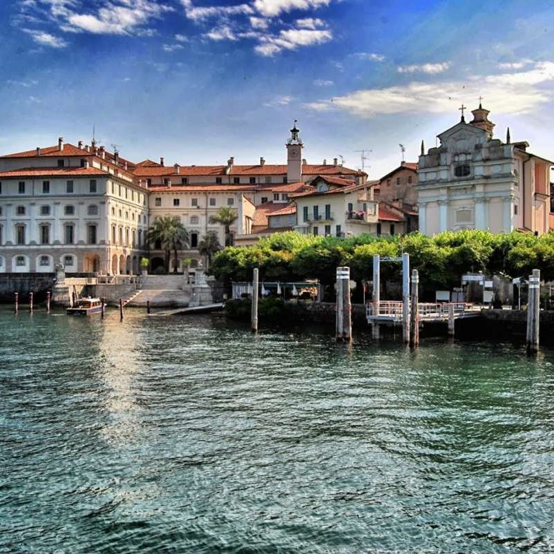 Vue du front de mer avec des bâtiments historiques aux toits de tuiles rouges, des arbres et des jetées le long d'un plan d'eau sous un ciel bleu partiellement nuageux.