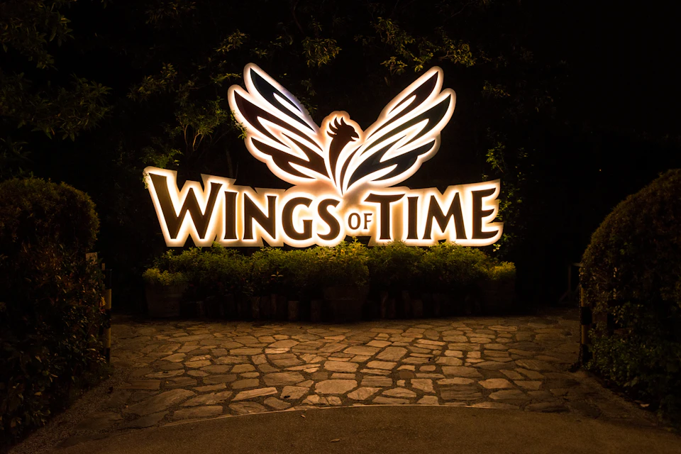Illuminated sign with the text "WINGS of TIME" and a winged design, set against a dark, tree-lined background on a stone pavement.