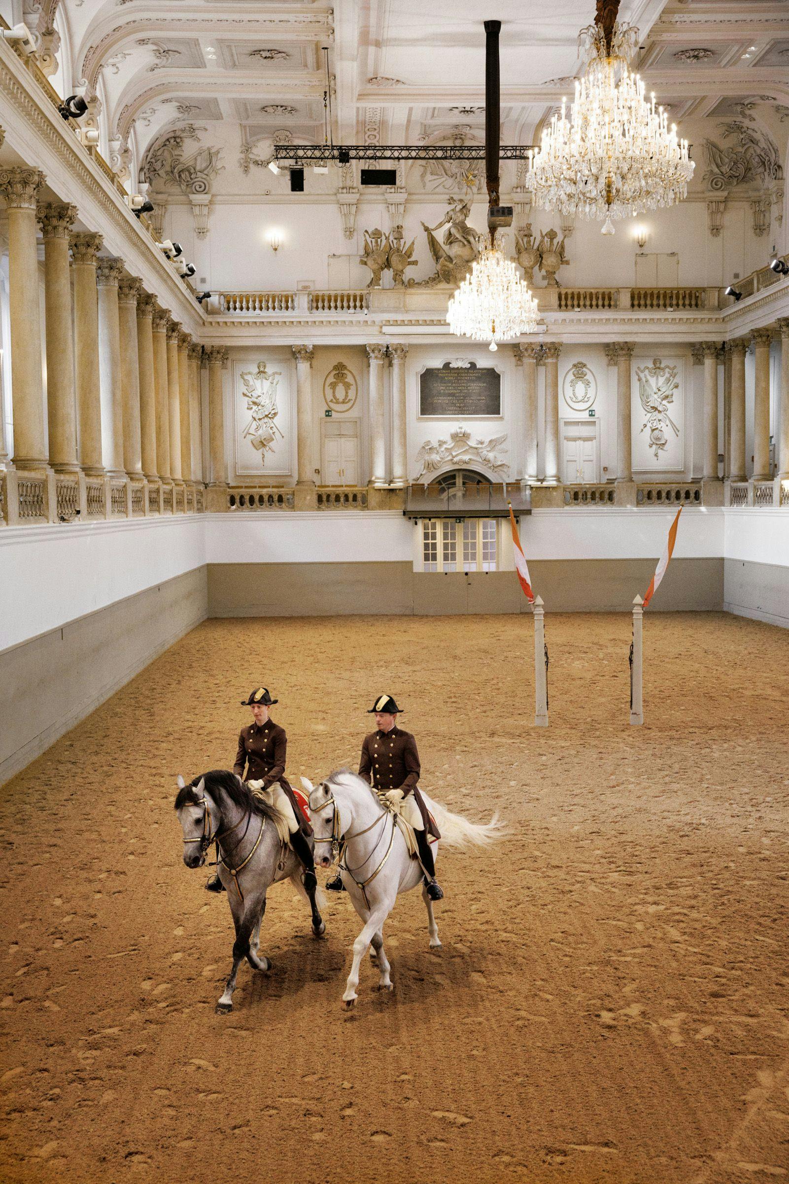 Two riders in formal attire on white horses in an ornate indoor arena with chandeliers and columns.
