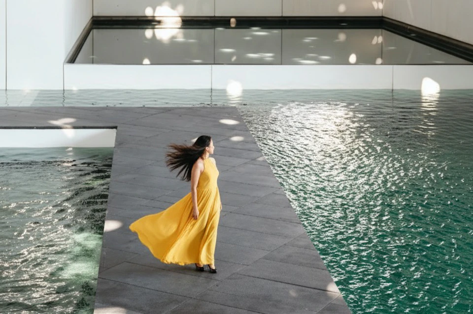 A woman in a flowing yellow dress stands on a gray tiled platform near a pool of water.
